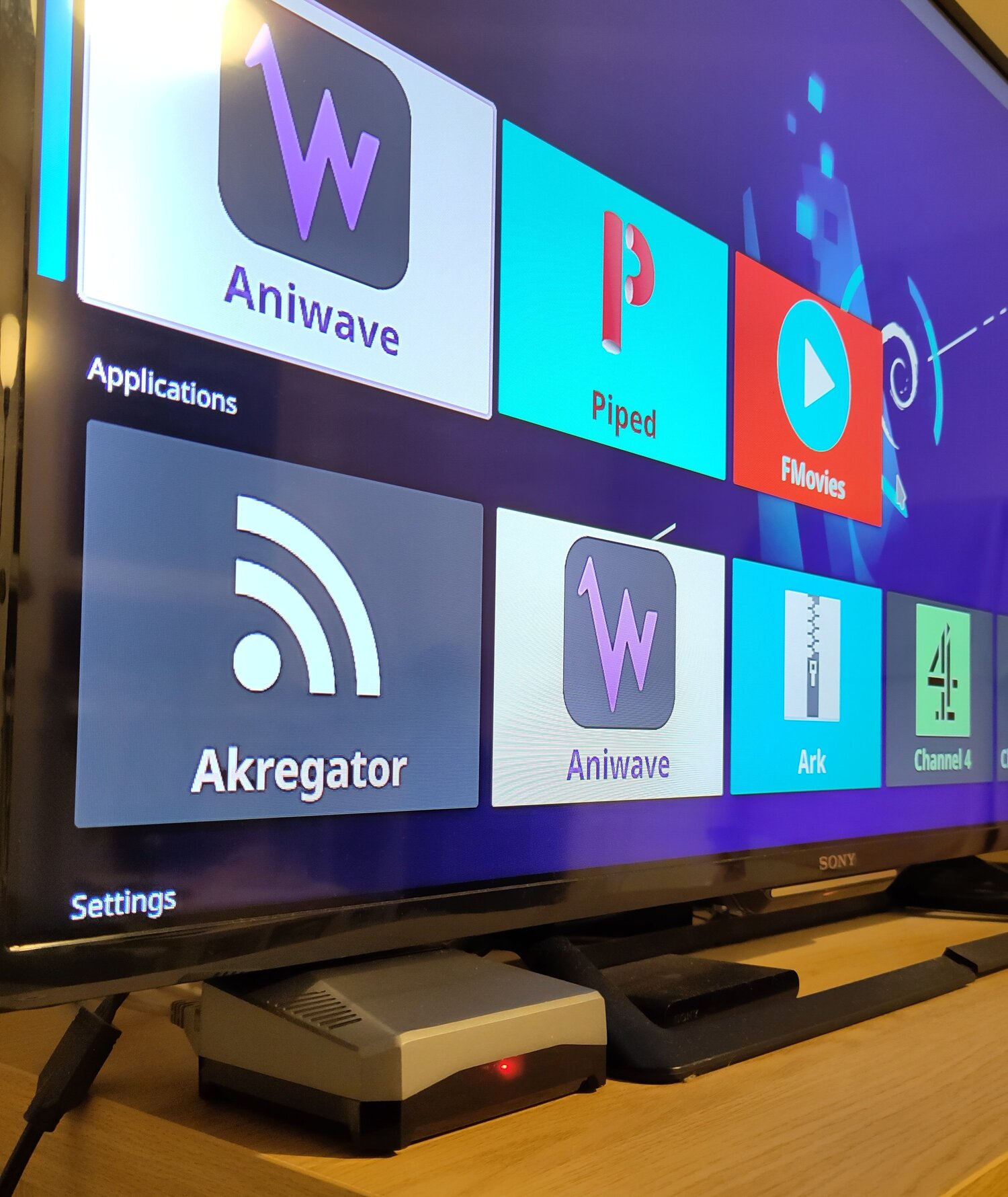 A Rspberry Pi 4b in an Argon One M.2. Sata case, powered
on and plugged into a TV that is on the KDE Plasma Bigscreen
home screen, with a Debian logo wallpaper.