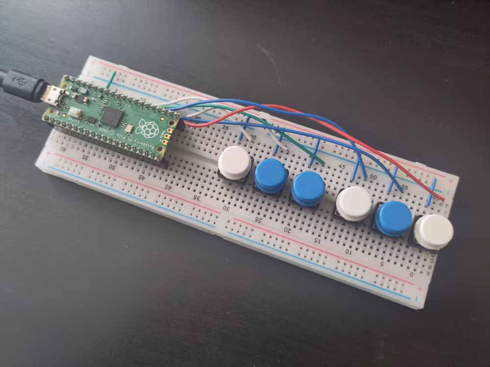 A Raspberry Pi Pico controller on a breadboard, wired
          up to six coloured buttons, in the pattern of white, blue,
          blue, white, blue, white.