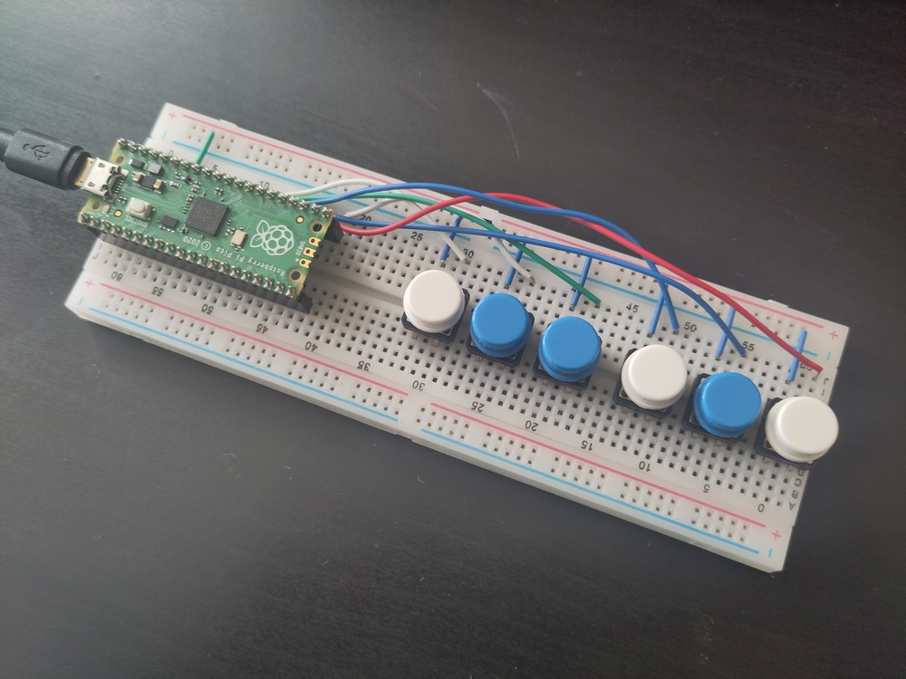 A Raspberry Pi Pico controller on a breadboard, wired
up to six coloured buttons, in the pattern of white, blue,
blue, white, blue, white.