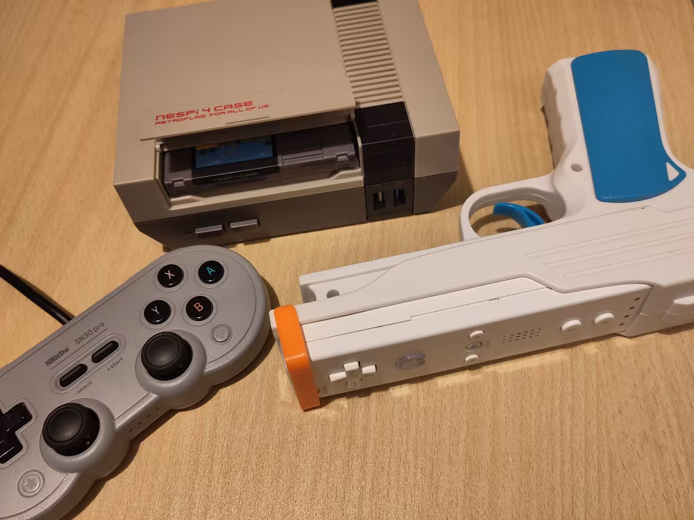 A NesPi-4 cased Raspberry Pi 4b, with the slot open
          to show a NES cartridge style SSD case, an 8BitDo SN30 Pro
          controller, and a WiiMote in a pistol lightgun adapter.