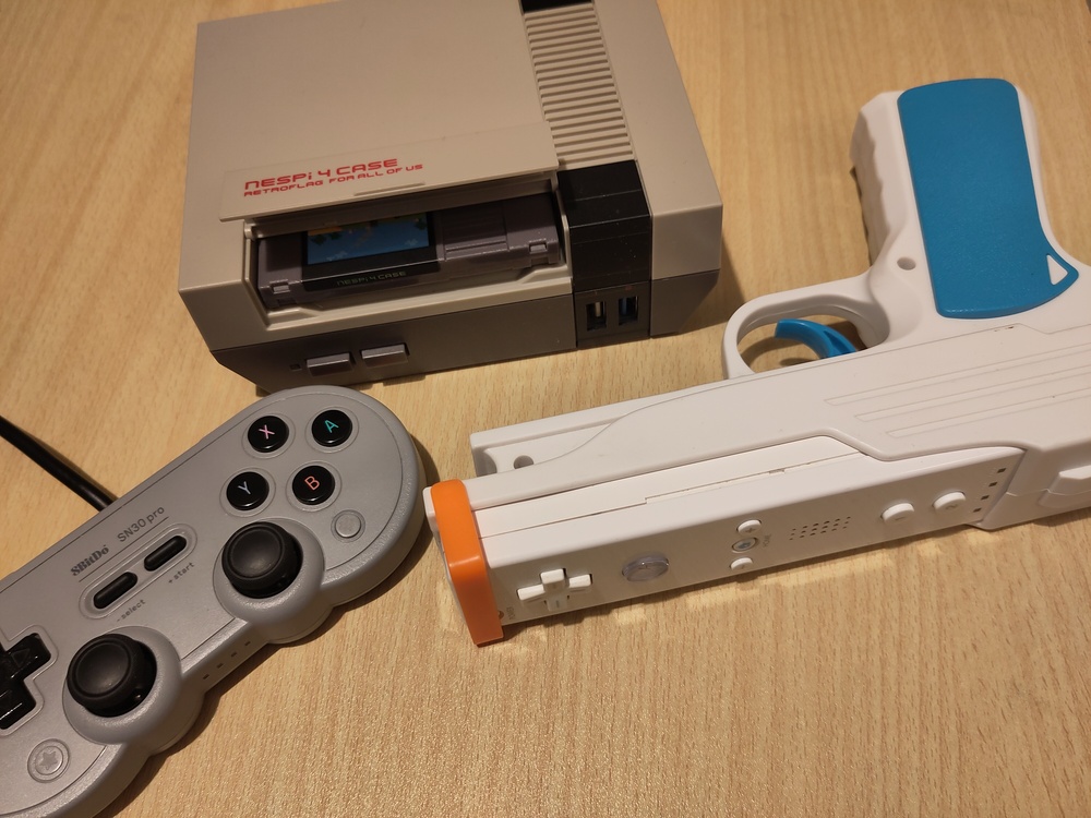 A NesPi-4 cased Raspberry Pi 4b, with the slot open
to show a NES cartridge style SSD case, an 8BitDo SN30 Pro
controller, and a WiiMote in a pistol lightgun adapter.